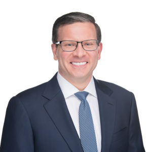 A person wearing a dark suit, white shirt, and blue tie is smiling against a white background.
