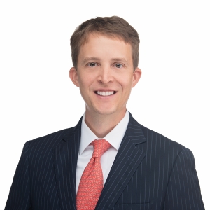 A man in a dark pinstripe suit and red tie smiles against a white background.