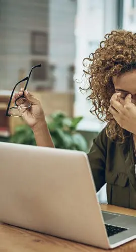 A person sitting at a desk with a laptop, holding their glasses and rubbing their eyes.