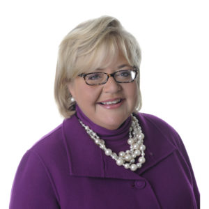 A smiling woman with short blonde hair, wearing glasses, a purple outfit, and a large pearl necklace is posed against a white background.