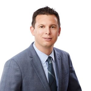 A person wearing a gray suit, light blue shirt, and matching tie poses for a professional portrait against a white background.