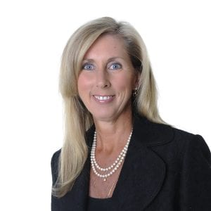 A smiling woman with long blonde hair wearing a black blazer and a pearl necklace against a white background.