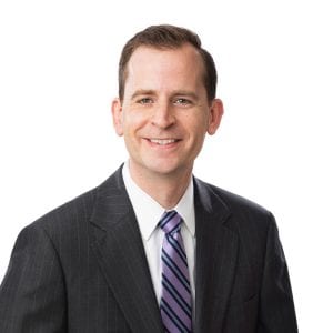 A smiling individual wearing a professional suit and a striped tie against a white background.