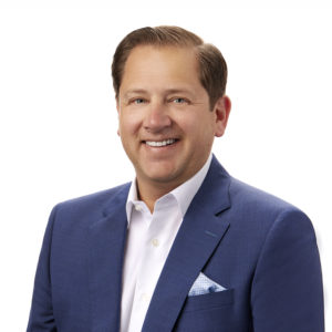 A man is smiling while dressed in a blue suit and white shirt against a plain white background.