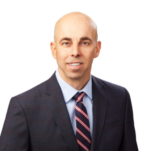 A bald person wearing a suit with a striped tie is smiling against a white background.