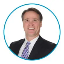 A professionally dressed individual in a business suit and tie is smiling against a white background, enclosed by a circular blue border.