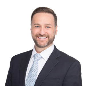 A person wearing a dark suit, white shirt, and a light blue patterned tie is smiling against a white background.
