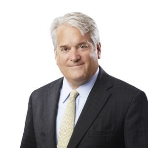 A man with white hair, wearing a dark suit, light blue shirt, and yellow tie is posing against a white background.