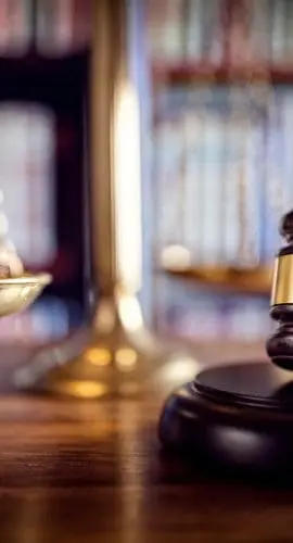 A wooden gavel and a brass scale symbolizing law and justice rest on a desk with books in the background.