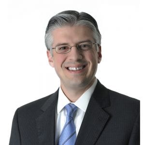 A smiling person with gray hair wearing glasses and dressed in a professional suit with a tie against a white background.