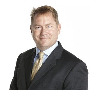 A man wearing a business suit with a tie is smiling at the camera against a white background.