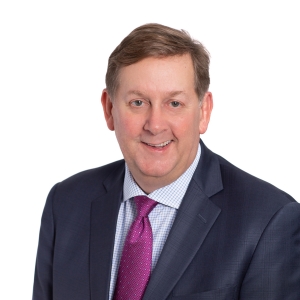 A man is wearing a dark suit jacket, a checkered shirt, and a pink tie, smiling against a plain background.