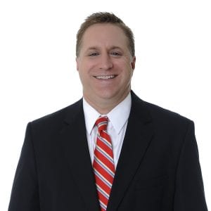 A man is smiling while wearing a black suit, white shirt, and a red striped tie against a white background.