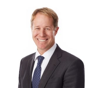 A smiling individual with short hair wearing a suit and a blue tie against a plain background.