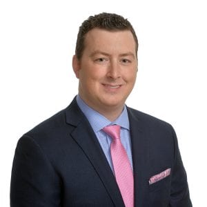A person is wearing a blue suit, a pink tie, and a matching pink pocket square, with a white shirt and a smile.