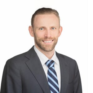 A man is wearing a suit and tie, smiling against a white background.